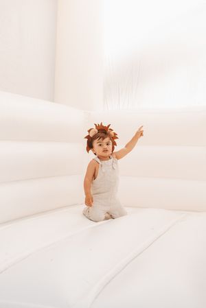 Playful toddler in white overalls and leaf crown poses on white backdrop.