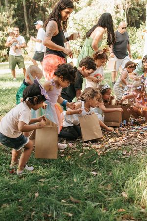 Children and adults bent over collecting treats from brown paper bags at an outdoor party.