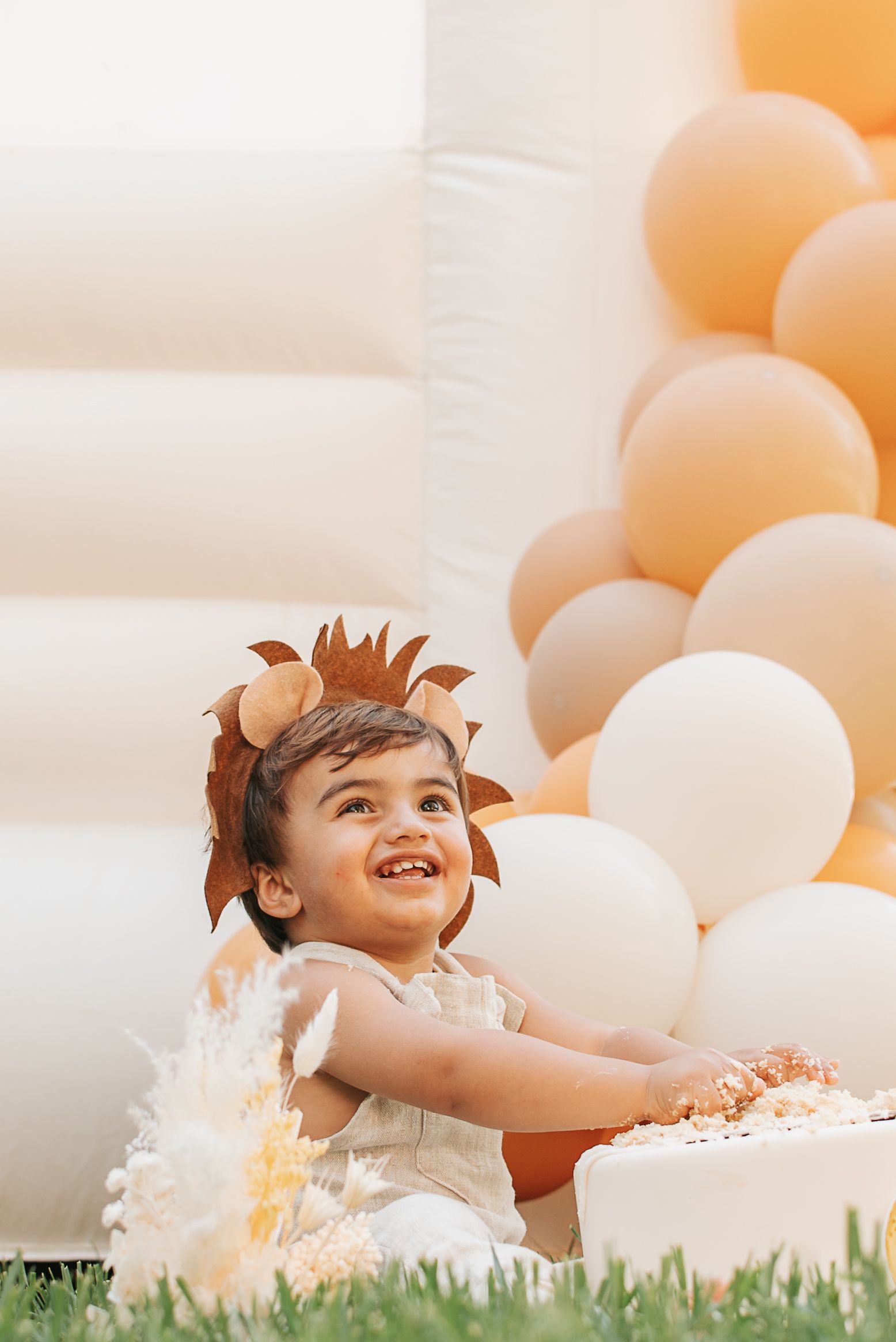 Child in lion costume smiles while playing with cake outdoors among peach and white balloons.