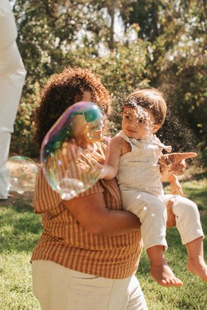 A joyful outdoor celebration with bubbles floating around as a toddler is held in the sunlit garden.