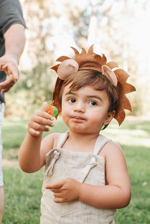 Child in linen overalls wearing lion headpiece holding small toy outdoors.