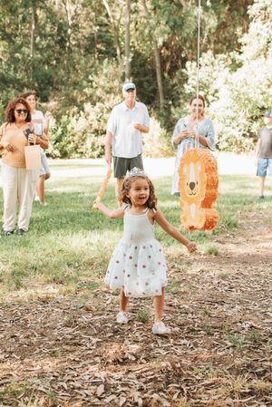 A child in a polka dot dress dances with an orange lion piñata at an outdoor celebration.
