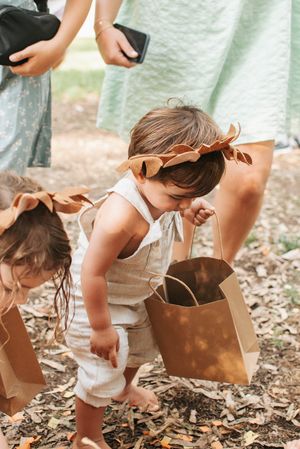 Small child in beige overalls with paper bag bending down at outdoor party.
