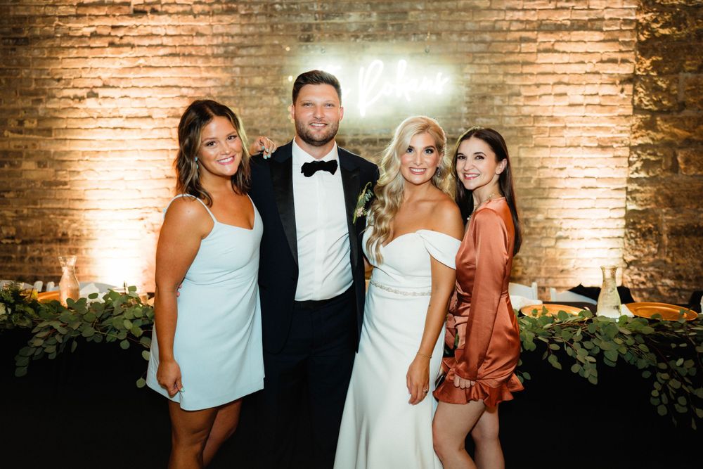 Wedding party poses against exposed brick wall with greenery decor at elegant reception.