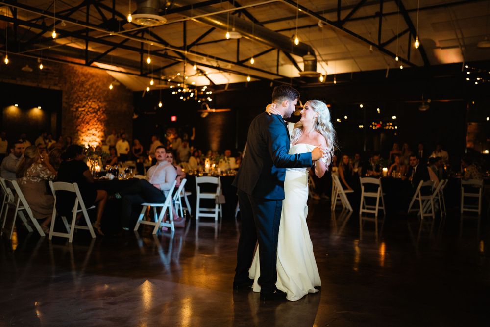 Romantic first dance in industrial wedding venue with uplighting and white chairs.