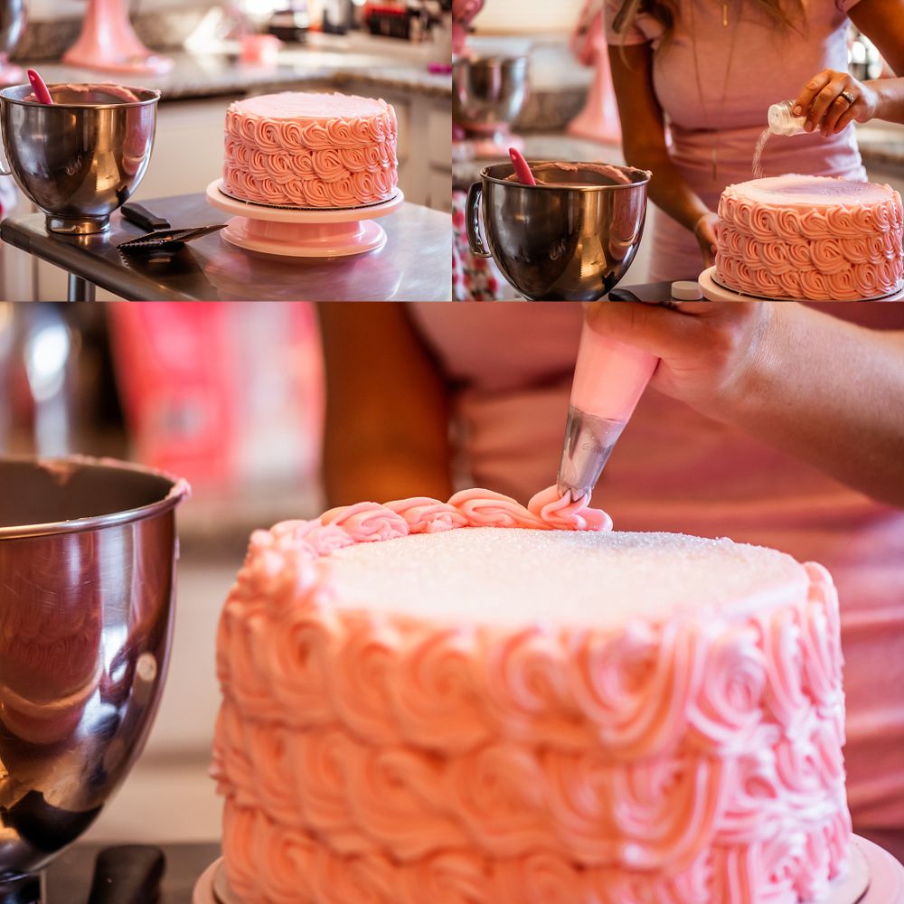 Pink frosted layer cake being decorated with piping tools and mixing bowls on counter.