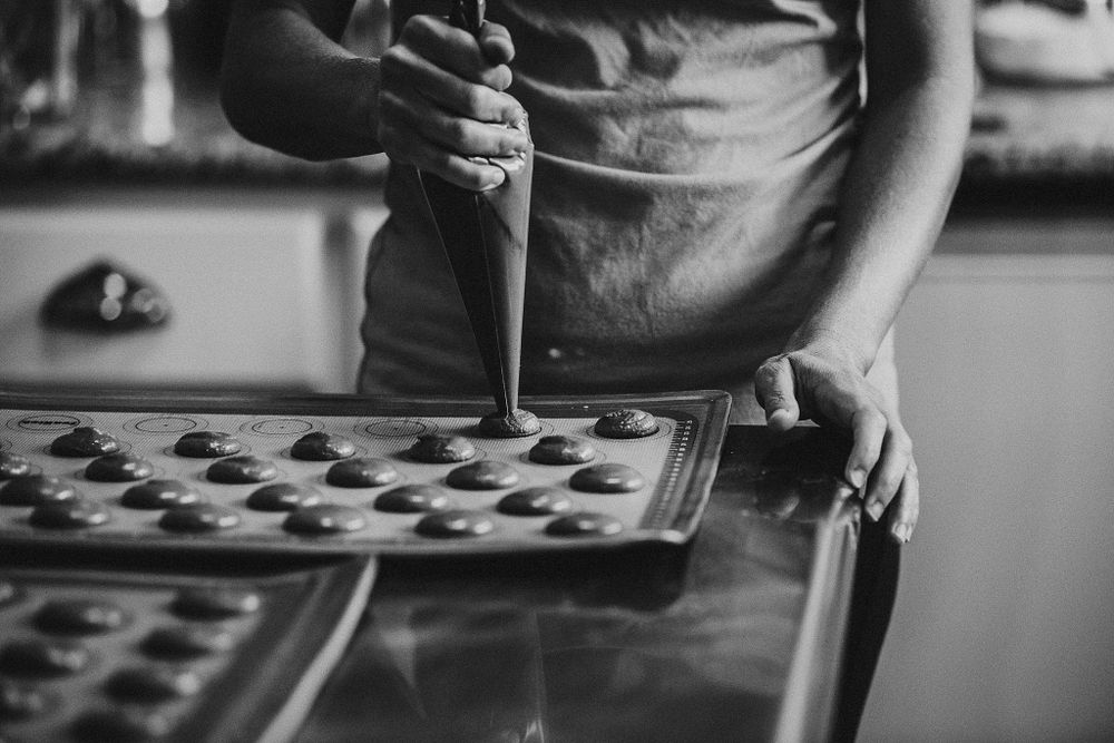 Black and white close-up of hands piping decorative elements onto baked goods.