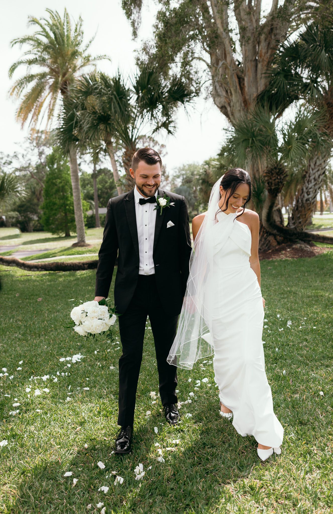 Newlyweds walking together on lush grass with palm trees and white bouquet in tropical setting.