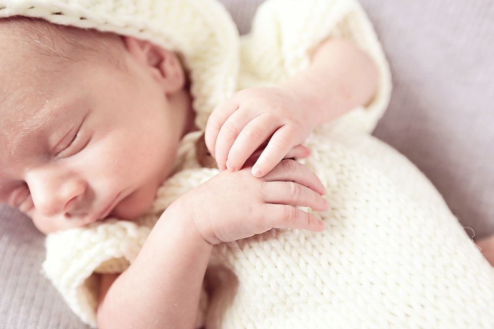 Sleeping newborn baby in cream knit outfit with tiny hands clasped together.