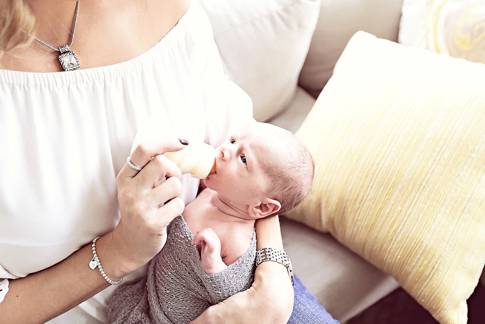 Mother bottle feeding newborn baby wrapped in gray blanket on white sofa.