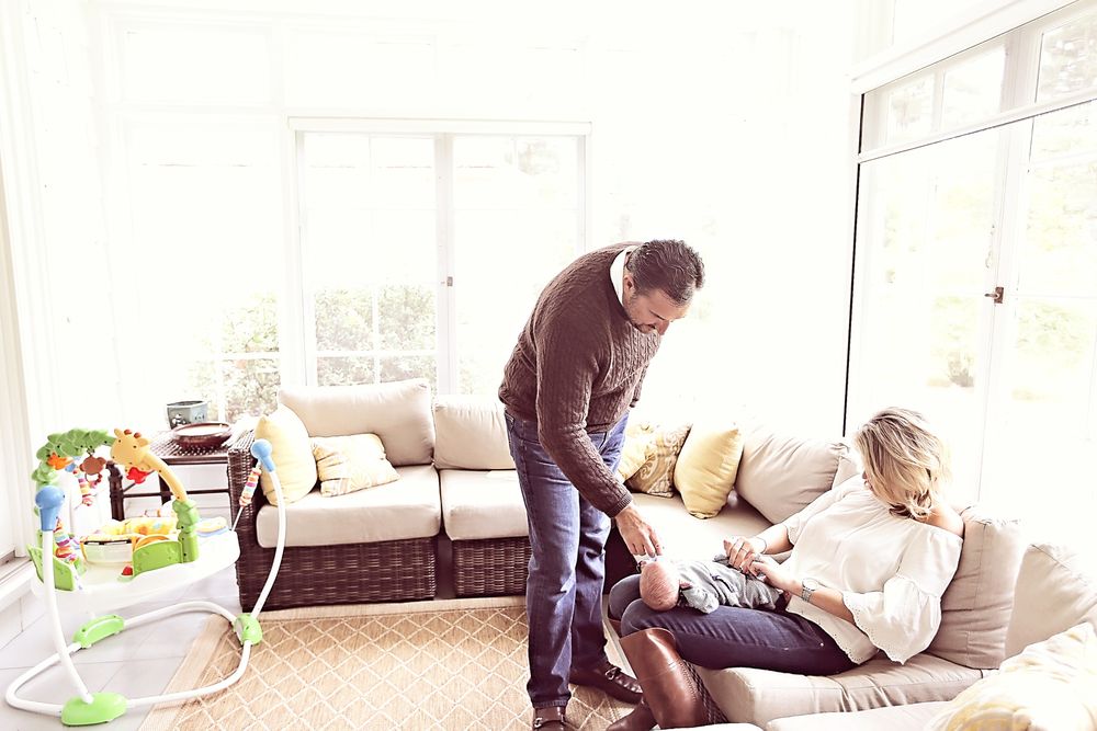A man leans over to interact with a woman holding a newborn baby on a cozy living room sofa.
