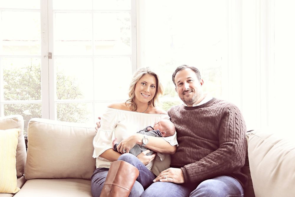 Happy couple sitting on a white sofa smiling while holding their newborn baby at home.