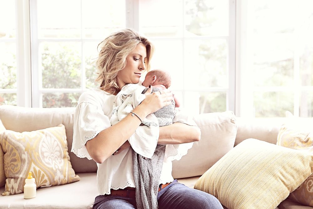 Blonde mother gazing lovingly at newborn baby in bright sunlit living room.