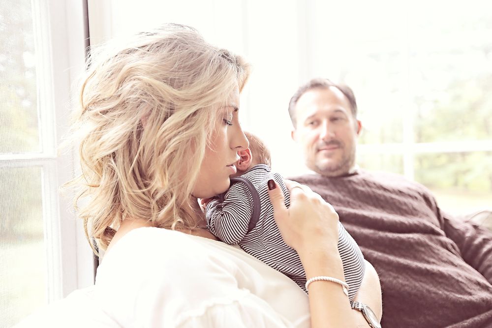 A blonde woman tenderly holds a newborn baby while a man smiles warmly in the background.