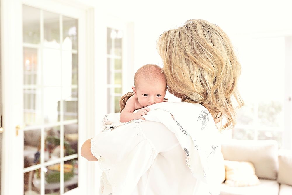 Mother holding newborn baby on her shoulder near bright white French doors.