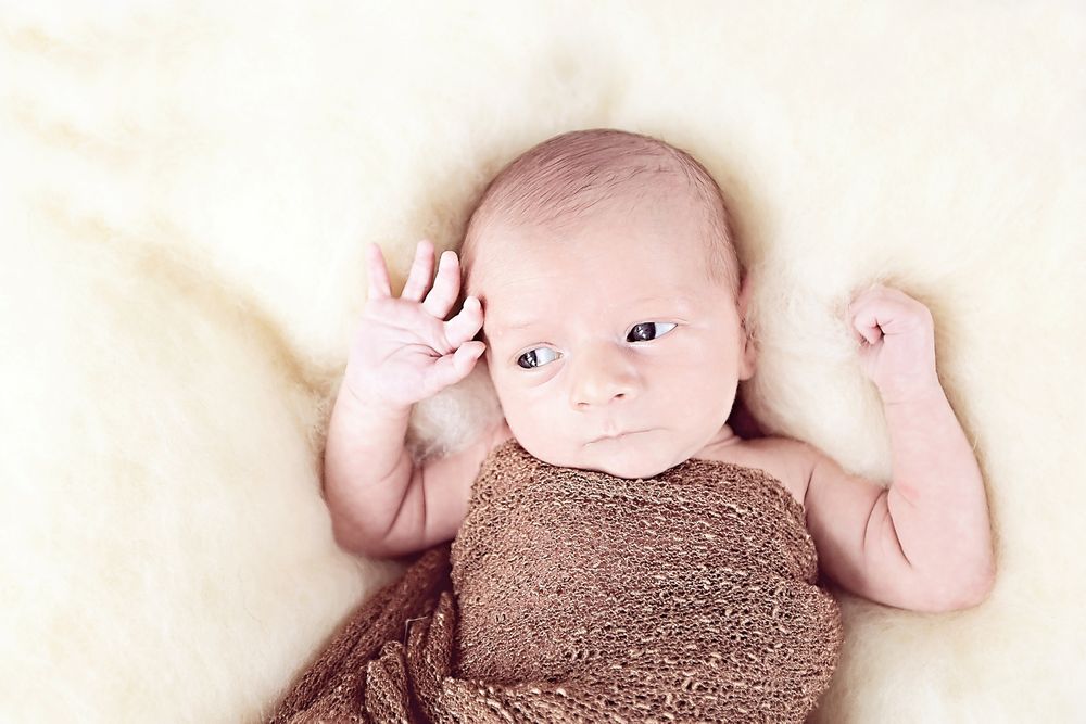 Awake newborn baby lying on white fur wrap with arms raised upward.