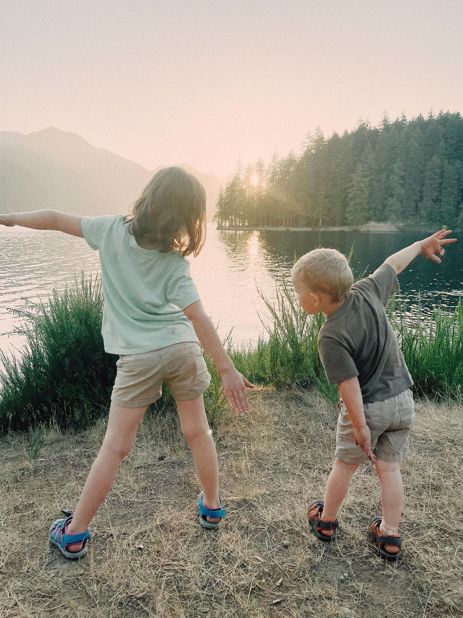Two small children bend down to explore something in the grass at sunset by a serene lake with mountains in the background.