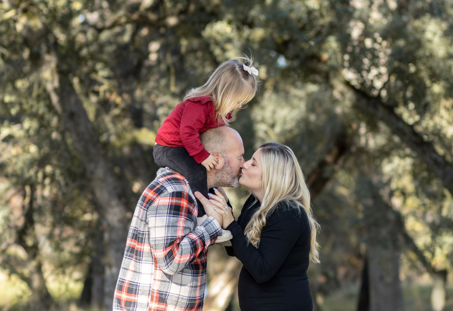 Parents kissing while lifting their young daughter above them outdoors among trees.