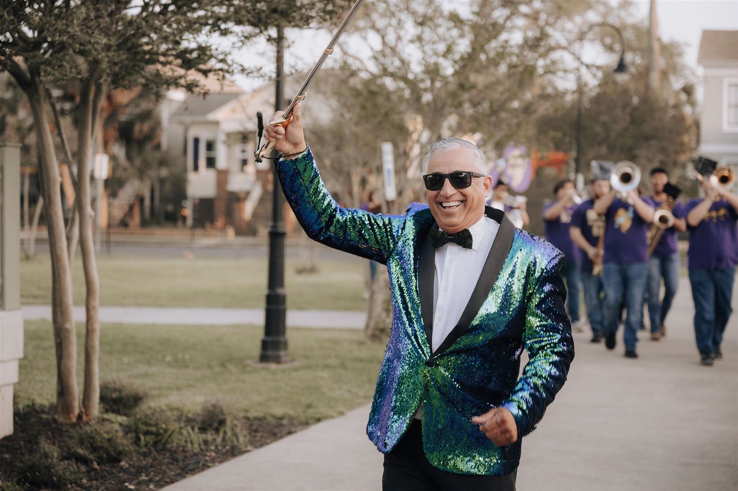 A person in a sparkly green jacket and bowtie leads a celebratory procession down a sidewalk.