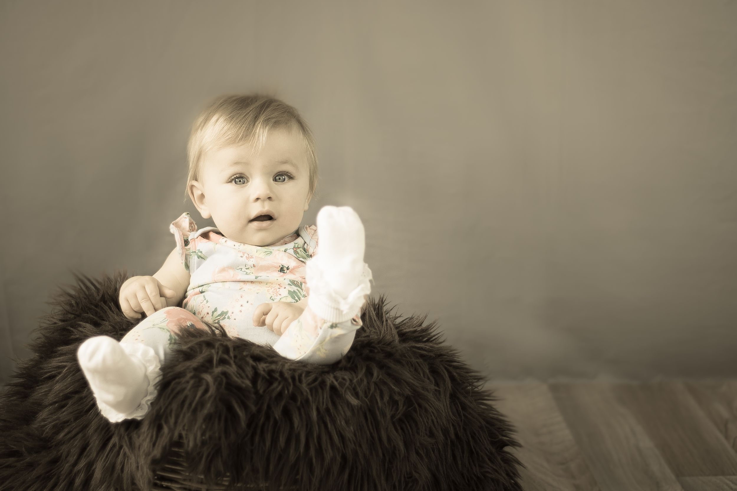 Baby photo session of baby In a sitting on a large fury cushion with white socks sonf patterned baby grow