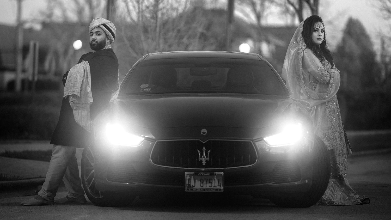 Couple leans on either side of a Maserati, the camera captivated by the glare of its headlights, embodying style, allure
