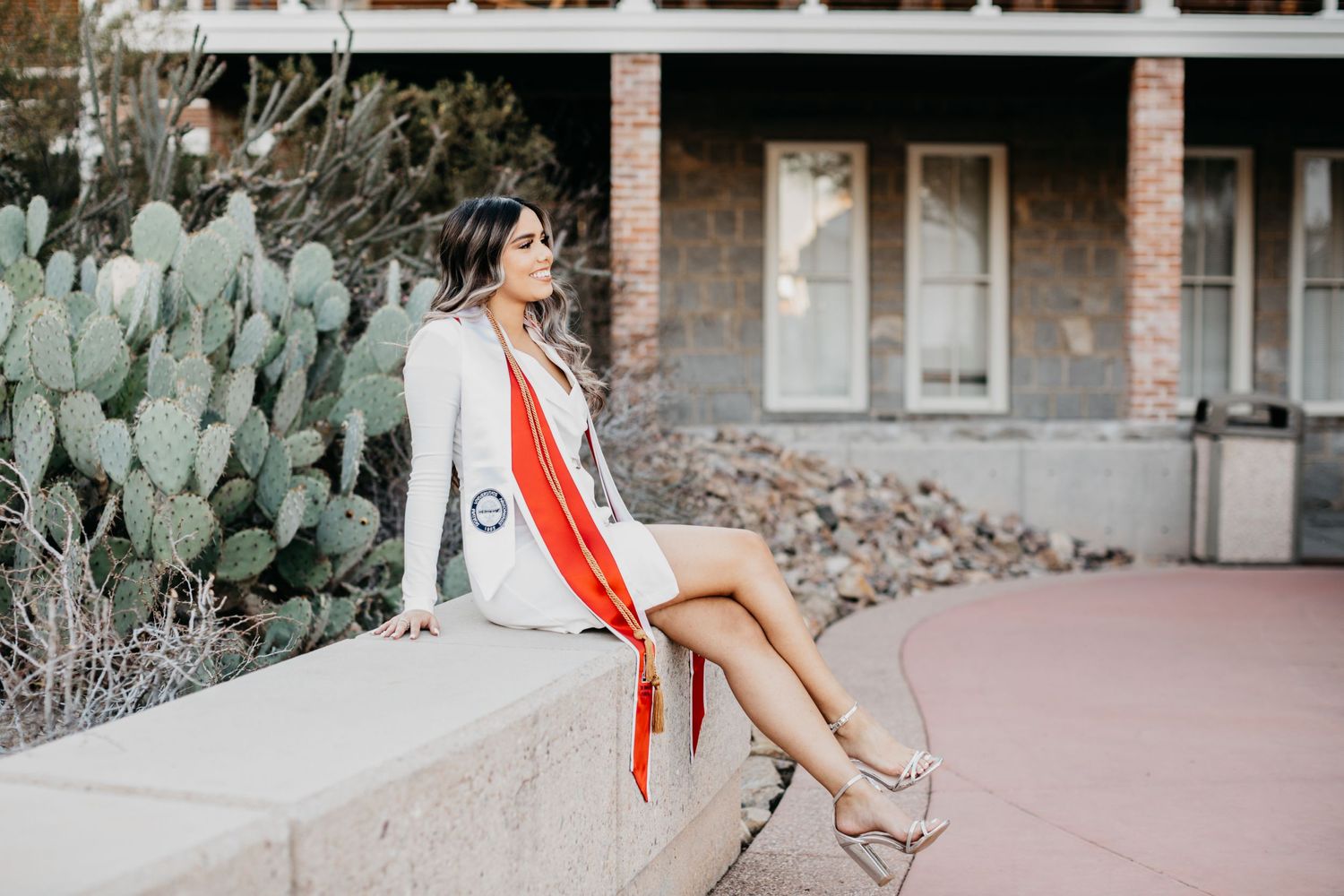 Tucson senior graduation portrait of a young woman sitting beside cactus plants wearing a red graduation stole.