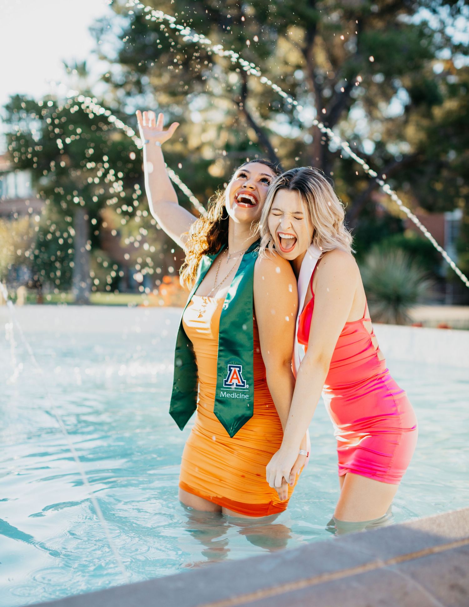 Tucson senior graduation photo of two girls celebrating by a fountain wearing colorful dresses and graduation stoles.