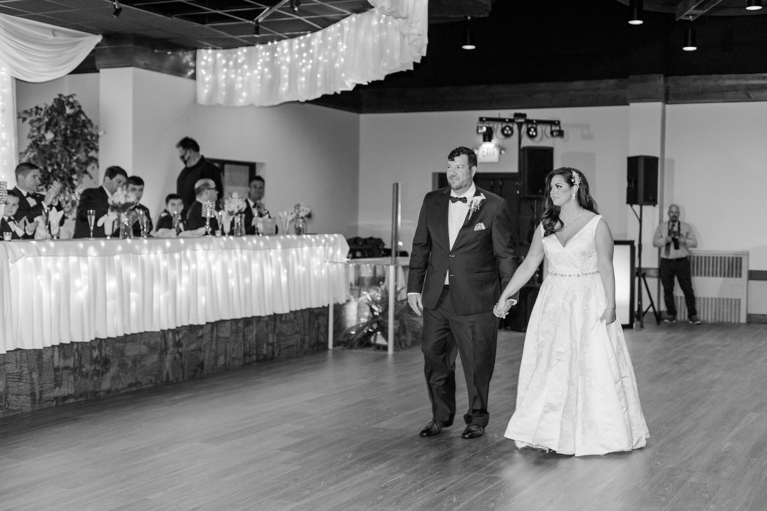 A black and white photo captures a couple walking across the dance floor at their wedding reception.