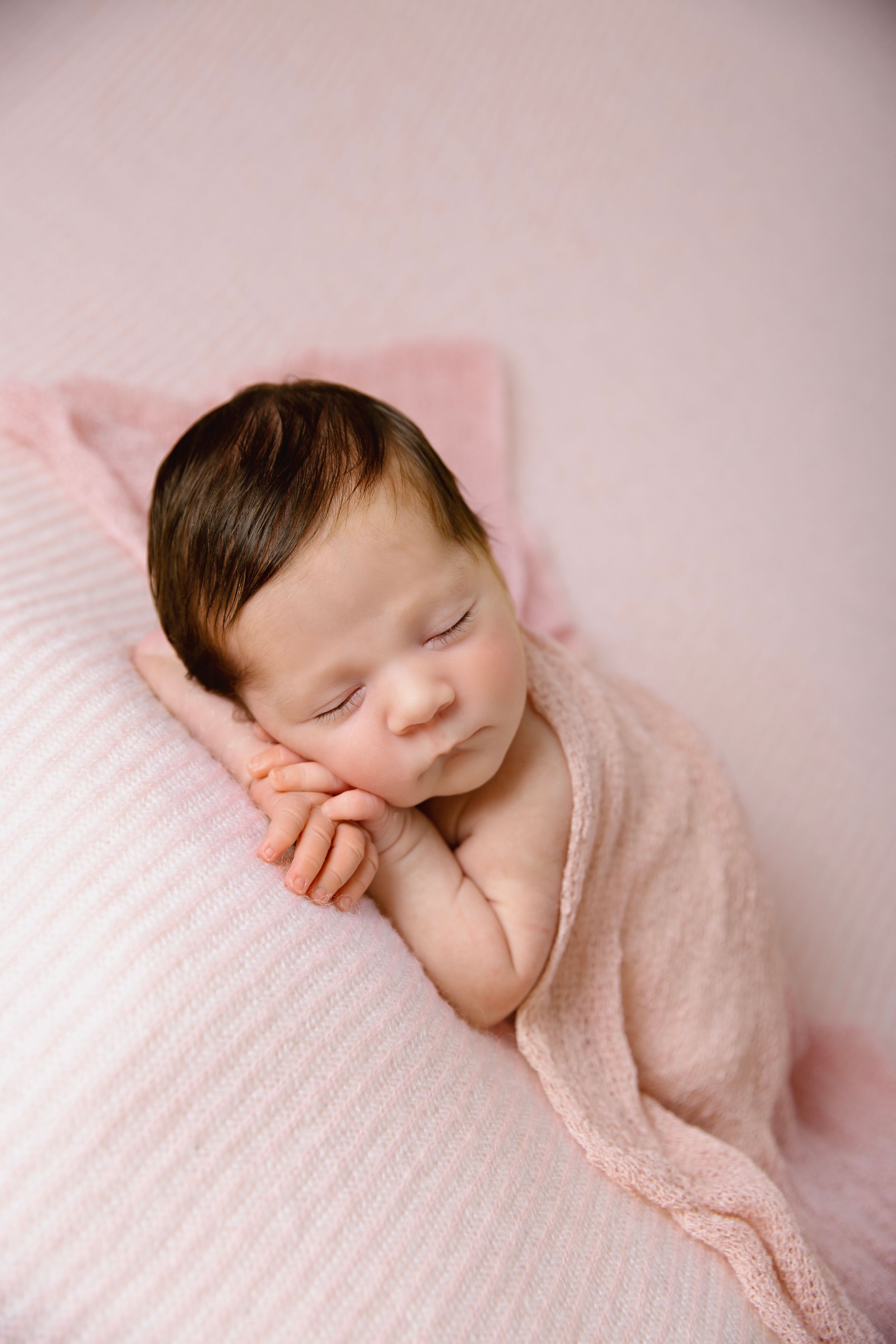Sleeping newborn wrapped in soft pink fabric on textured background.