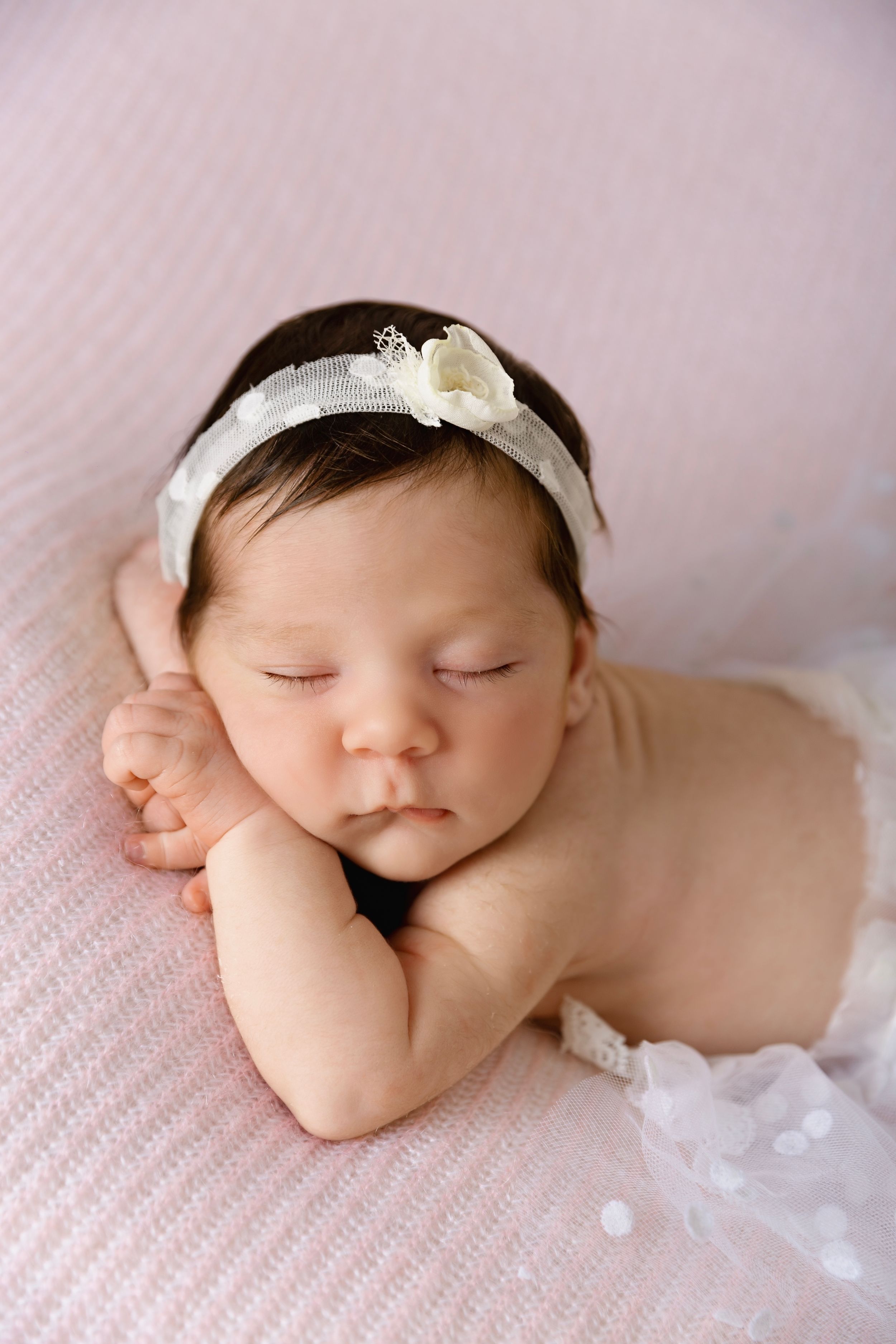 Peaceful sleeping newborn wearing white headband on pink textured background.