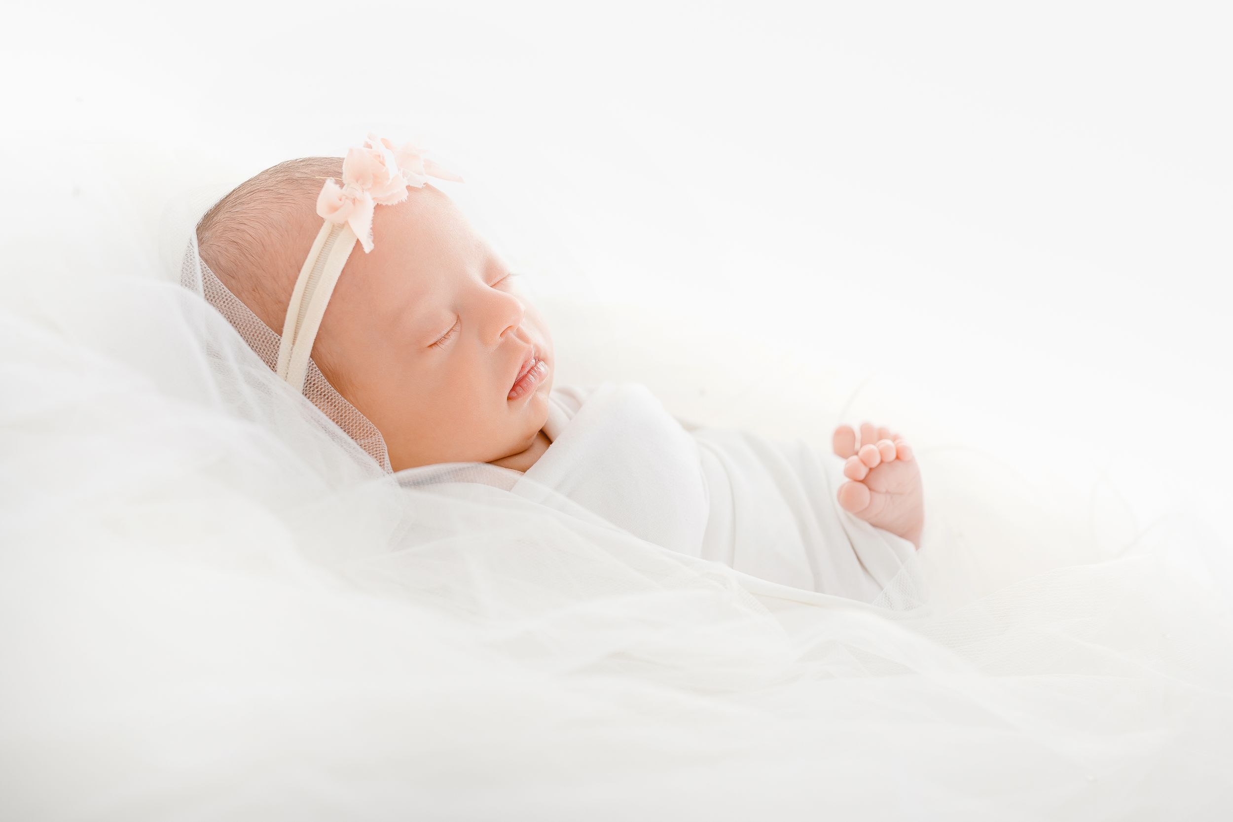 Serene newborn portrait with white bow headband against soft white background.