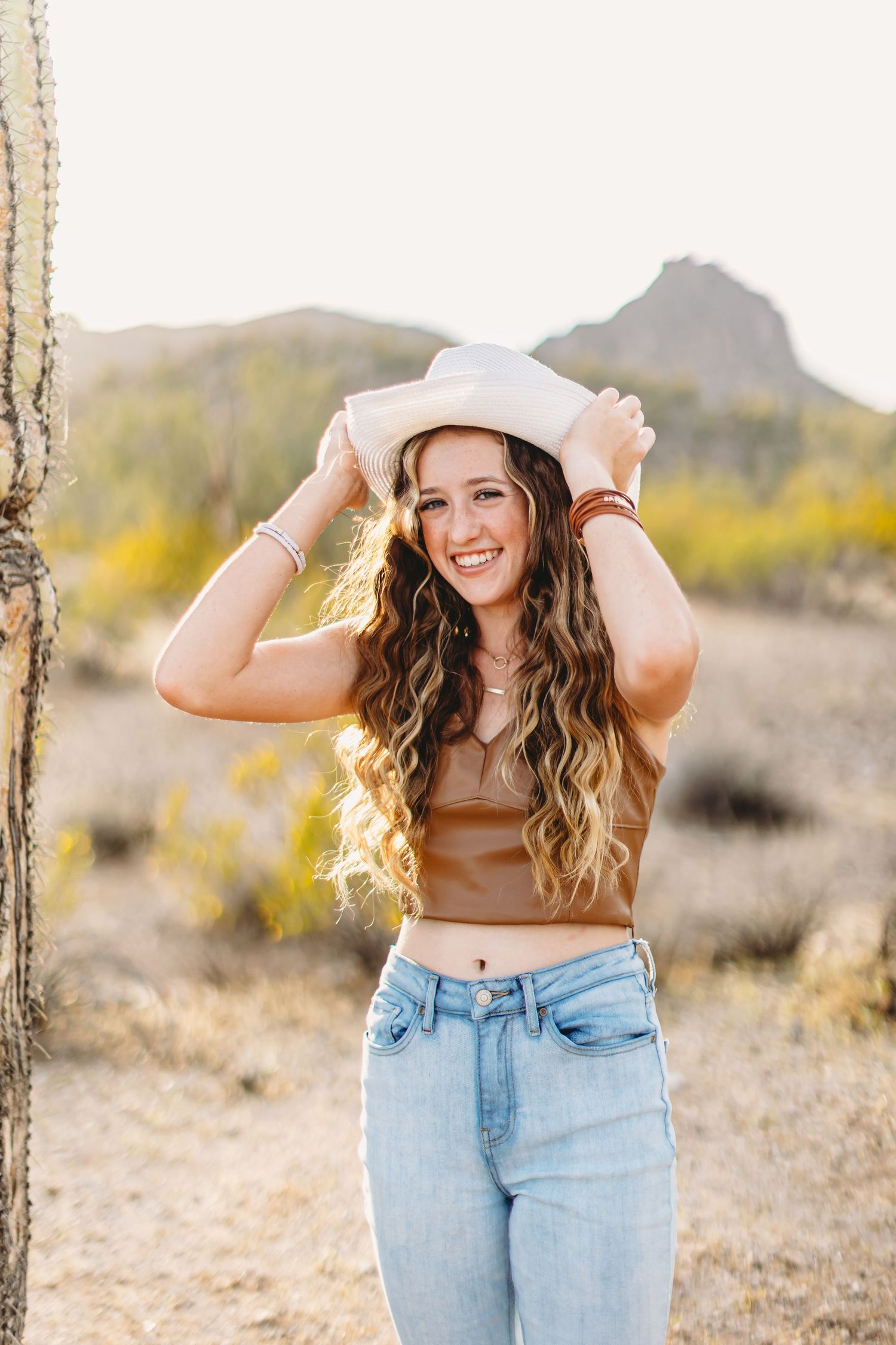 Woman in western hat and jeans poses next to saguaro cactus in desert landscape.