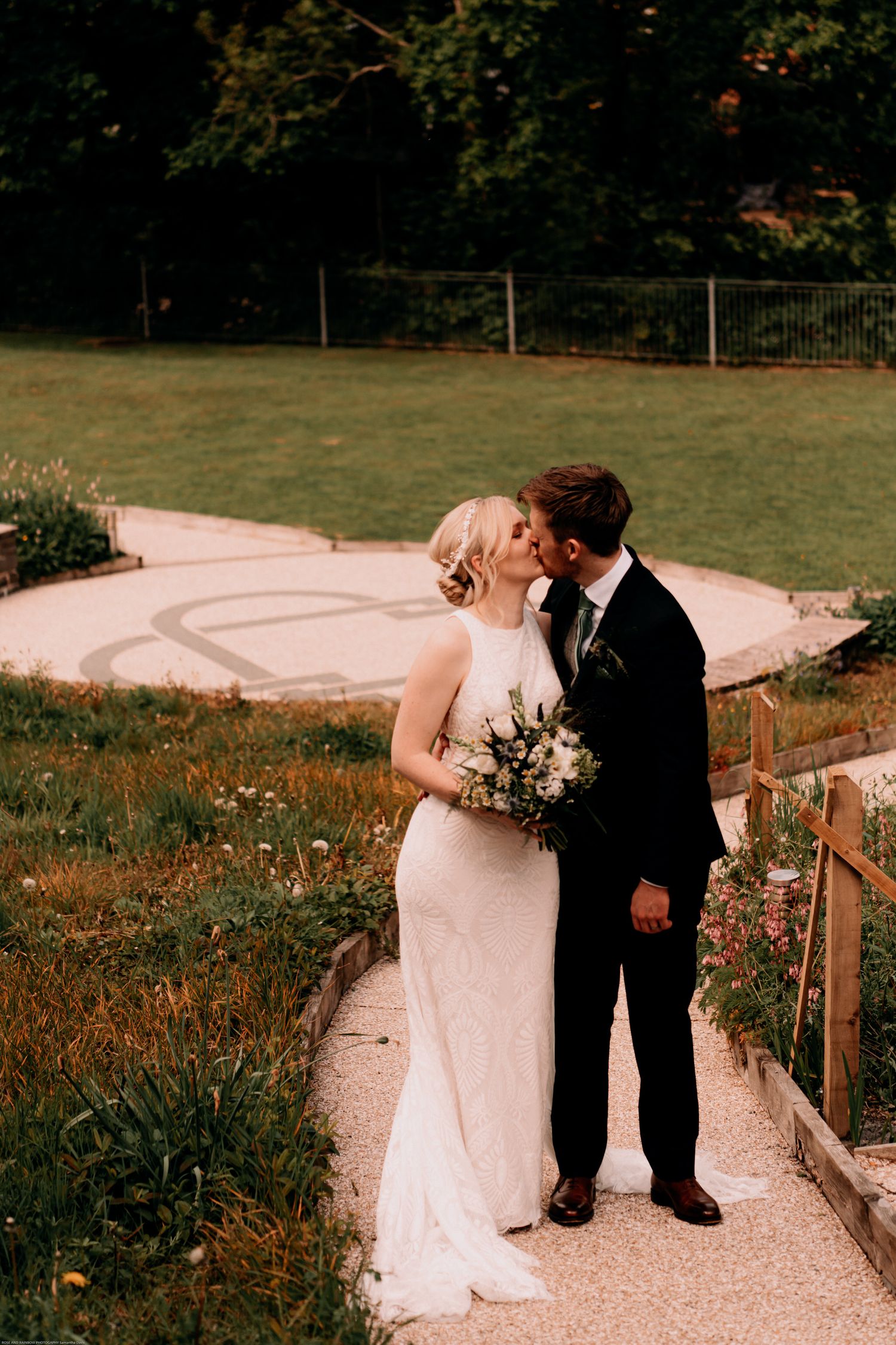 A couple shares a romantic moment on a garden path while holding a wedding bouquet in an outdoor setting at sunset.