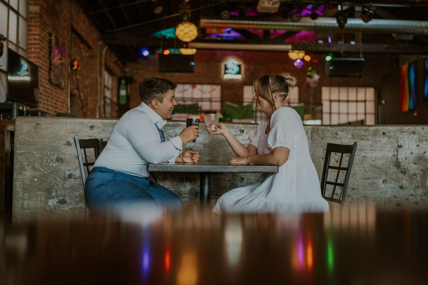 Couple enjoying drinks at a rustic bar with colorful lighting in the background.
