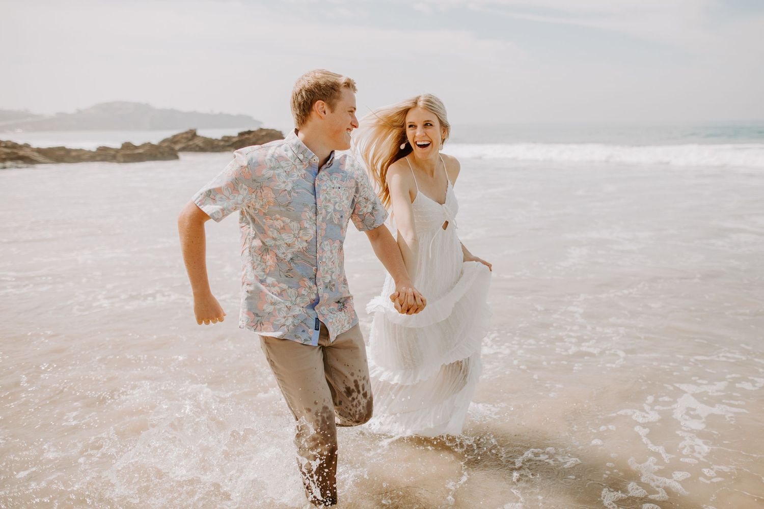 A couple holding hands and running joyfully through the ocean waves on a misty beach.