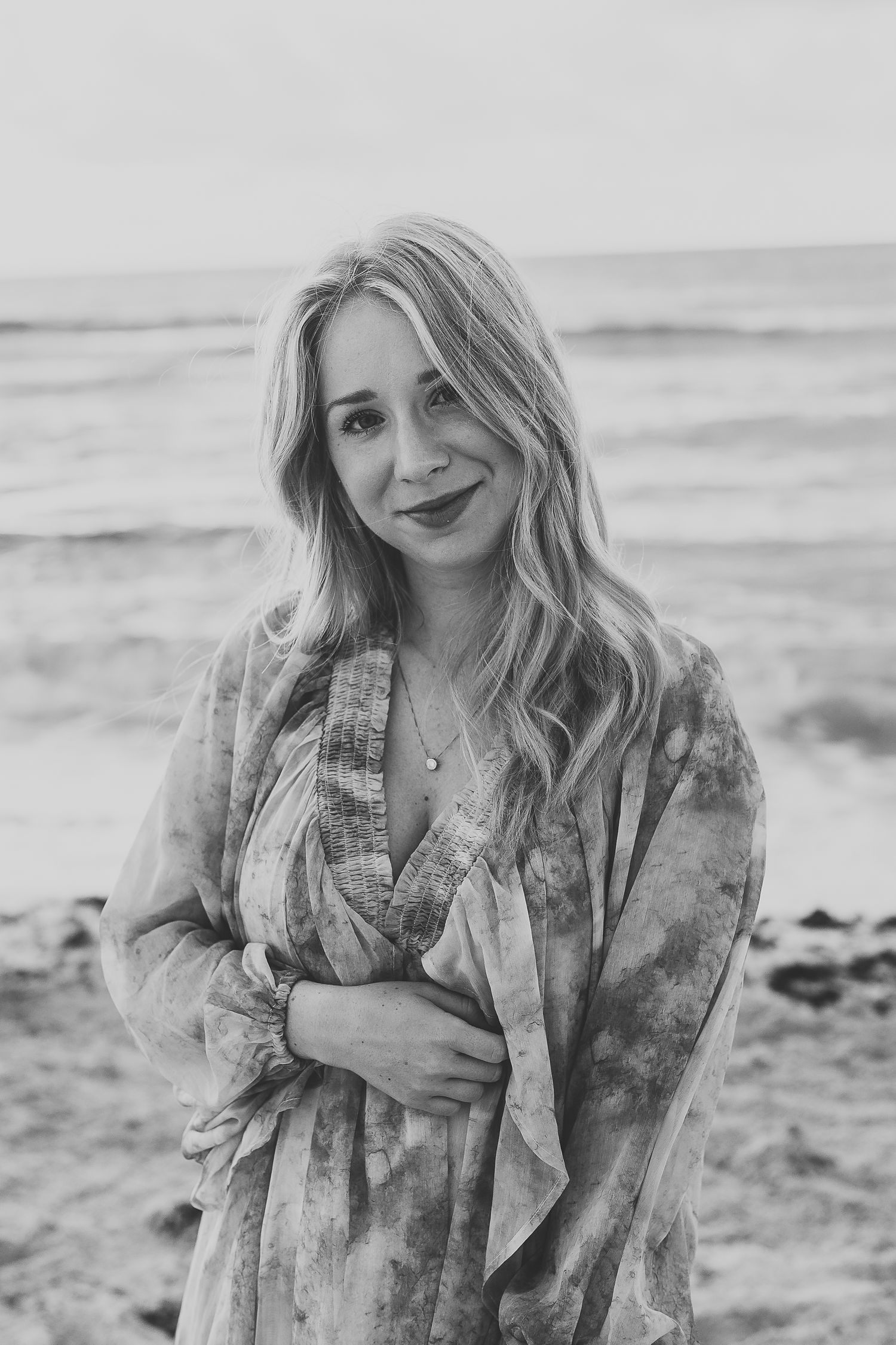Woman in flowing dress stands on beach shore in black and white portrait.