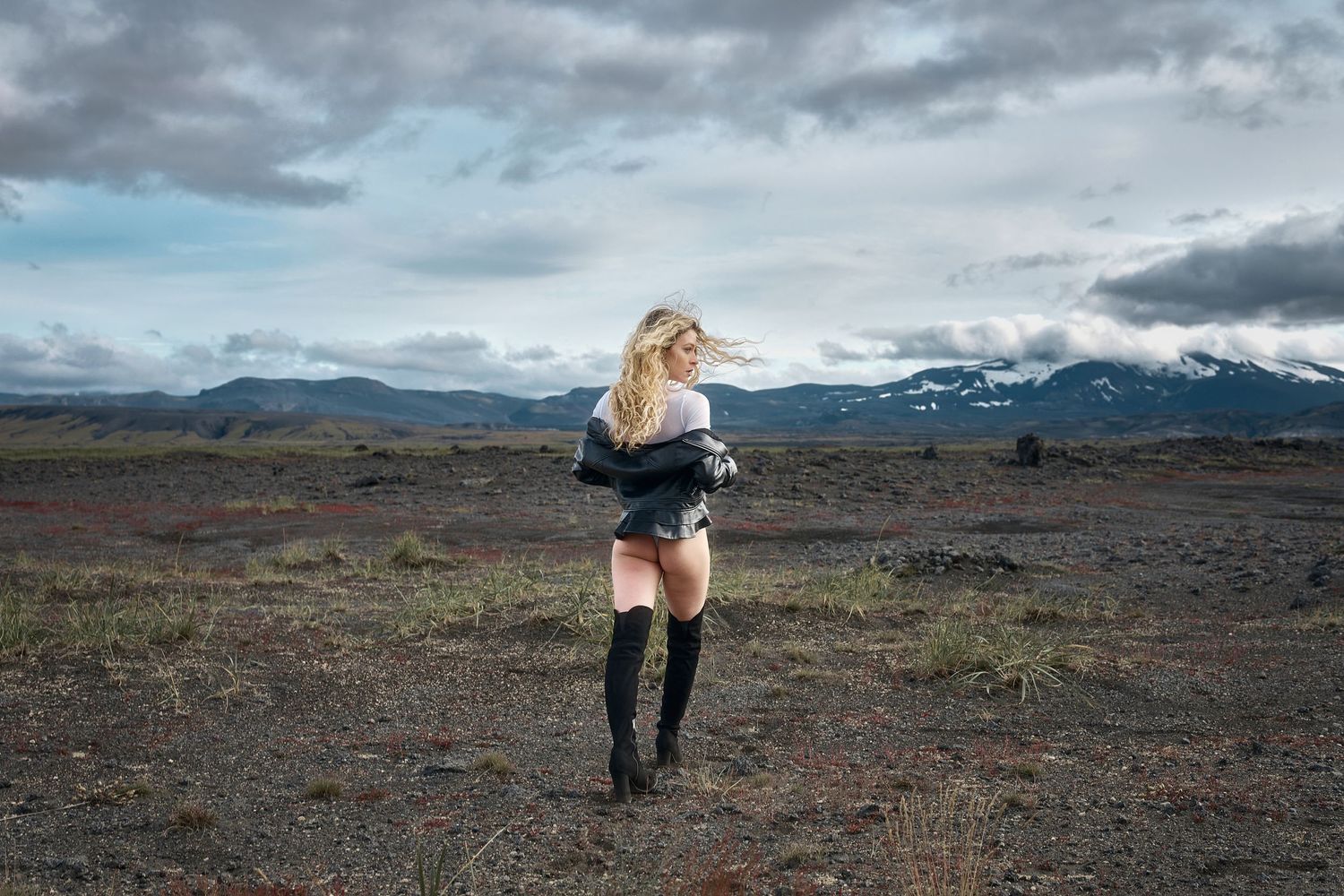 Figure in thigh-high boots stands against dramatic mountain landscape under cloudy skies.