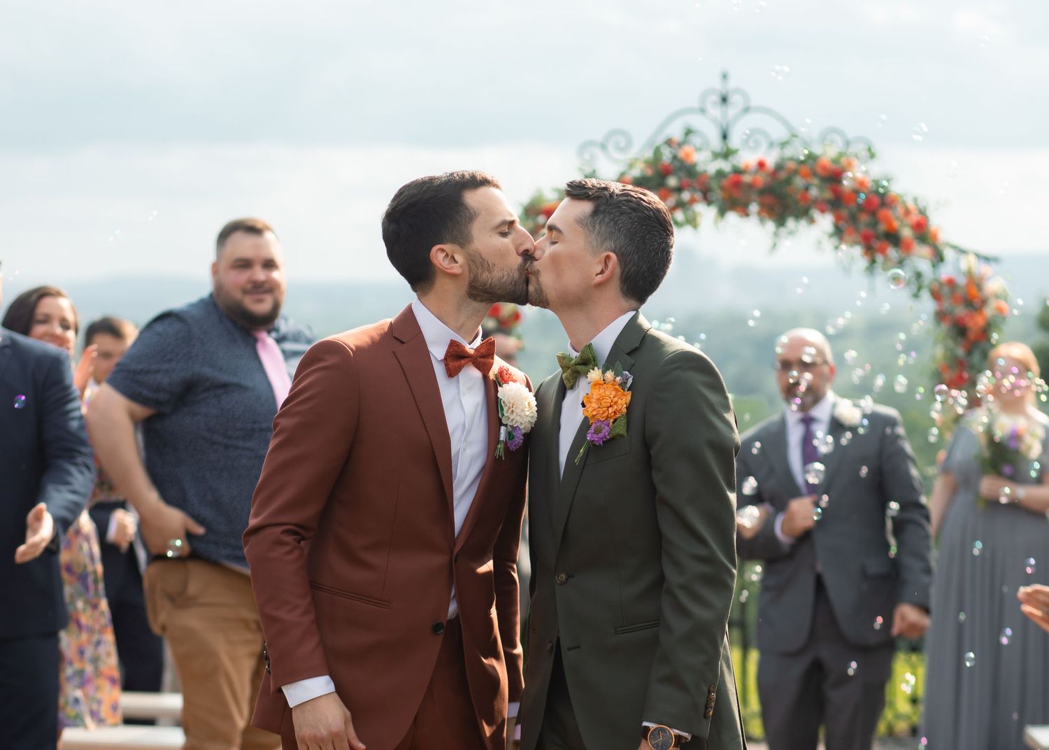 Two men in formal attire share a romantic kiss under a floral arch during their outdoor wedding ceremony in Connecticut, USA.