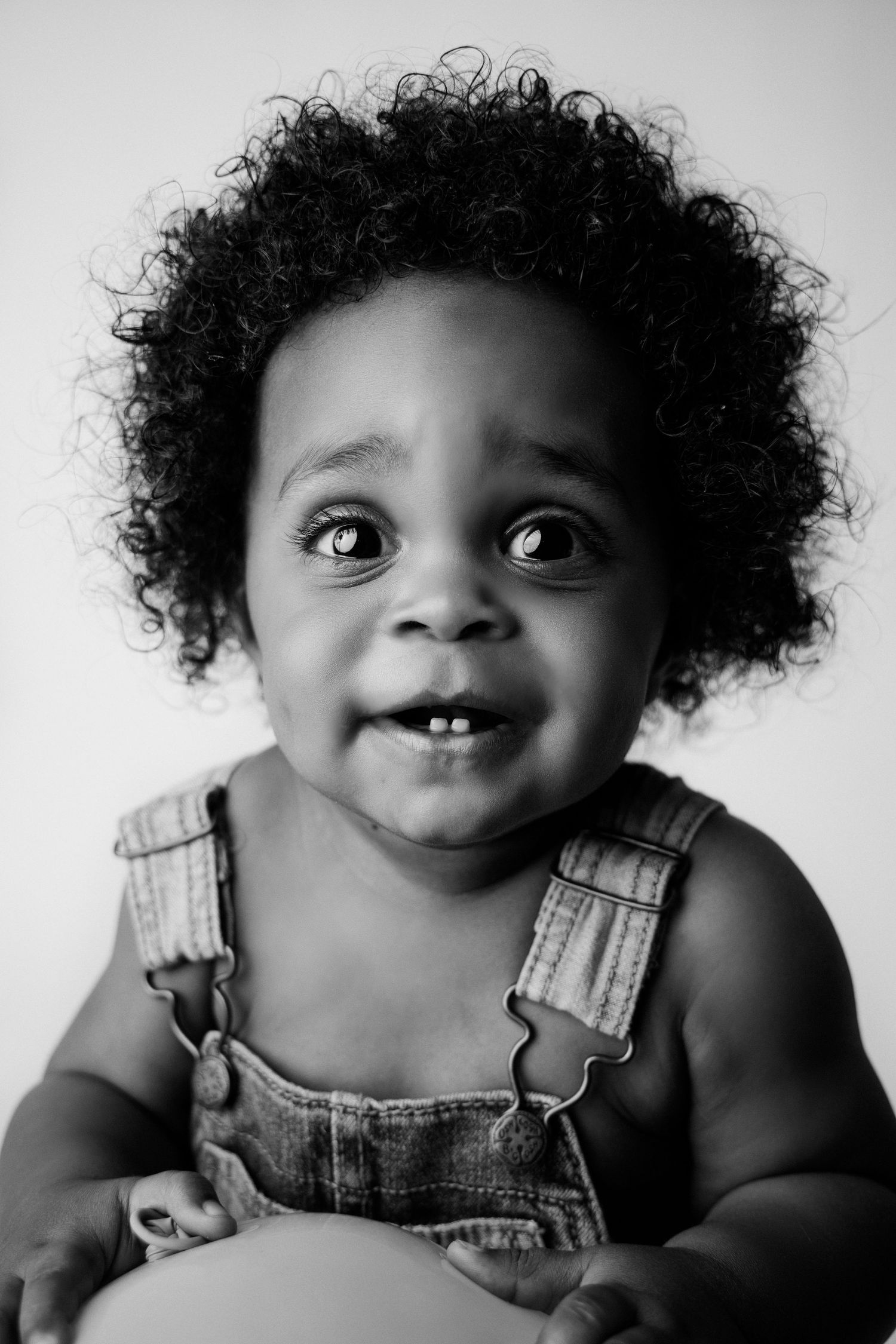 Black and white portrait of toddler in overalls with natural curly hair.