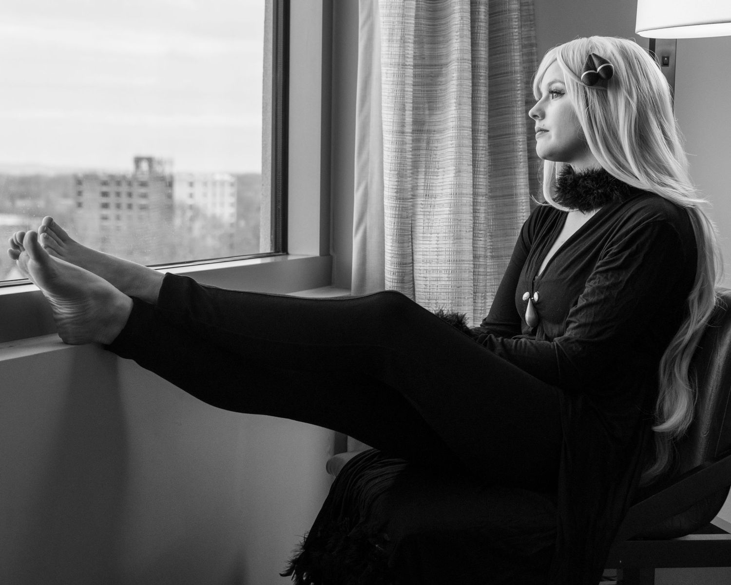 A person in black clothing relaxes by a window with feet up in dramatic black and white.
