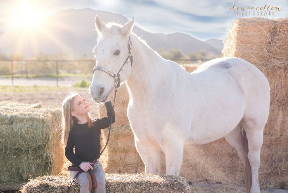 Young girl kisses white horse near hay bales with mountains in golden sunset light.