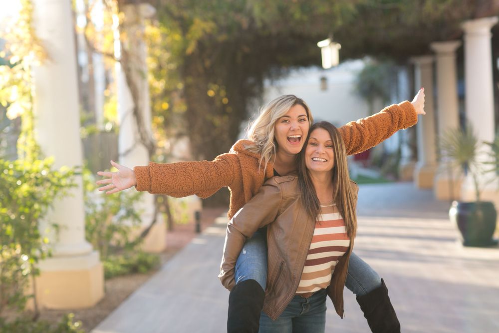 Two young women laugh joyfully as one gives the other a piggyback ride on a sunny tree-lined path.