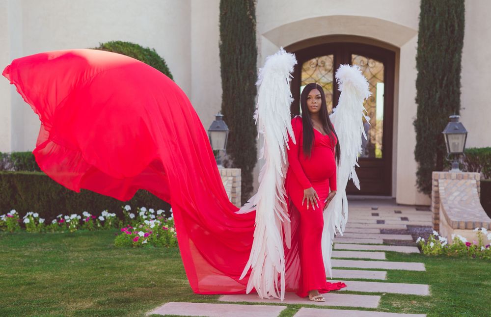 Woman in flowing red gown with white angel wings stands before elegant mansion entrance.