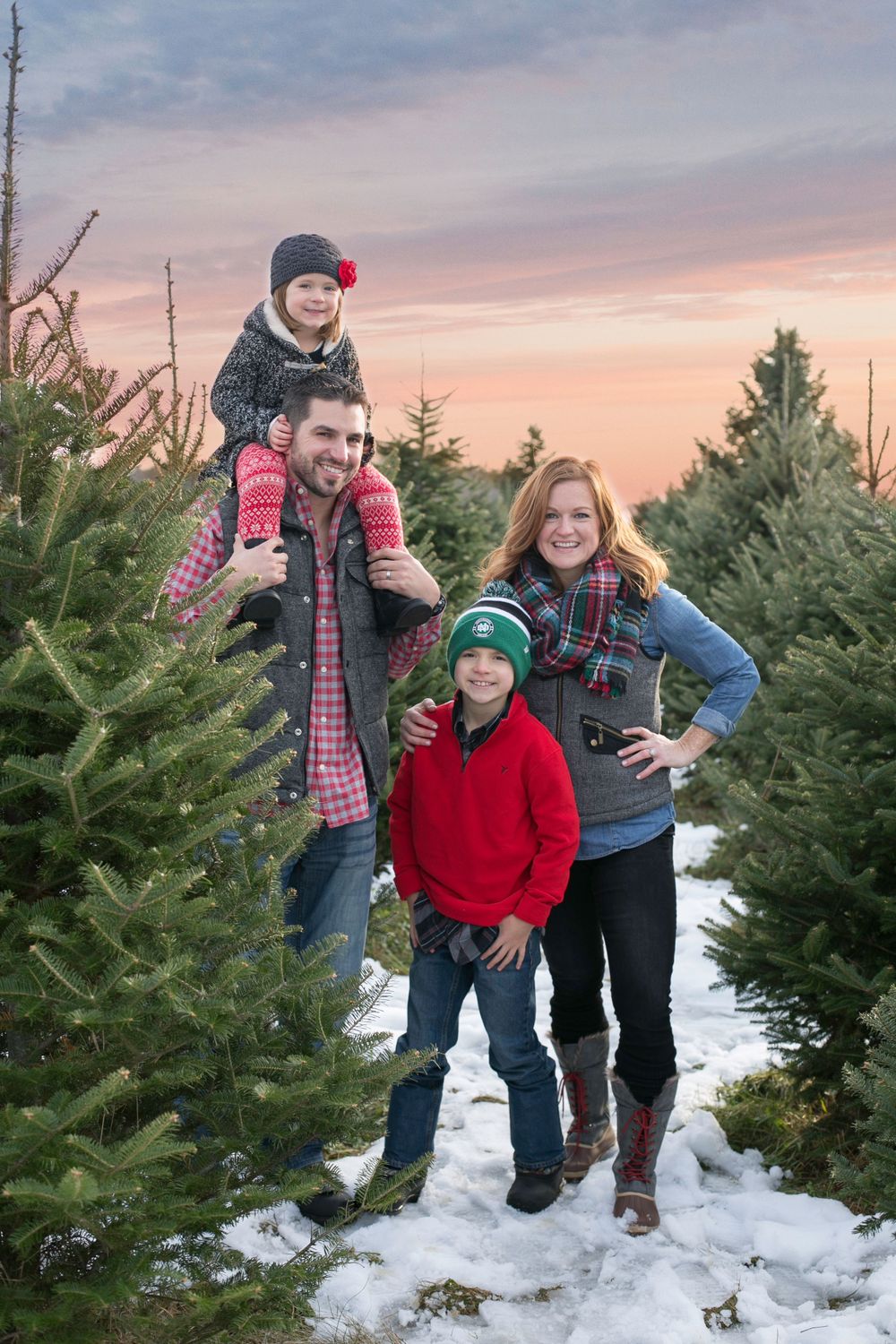 Happy family of four posing at Christmas tree farm during colorful winter sunset.