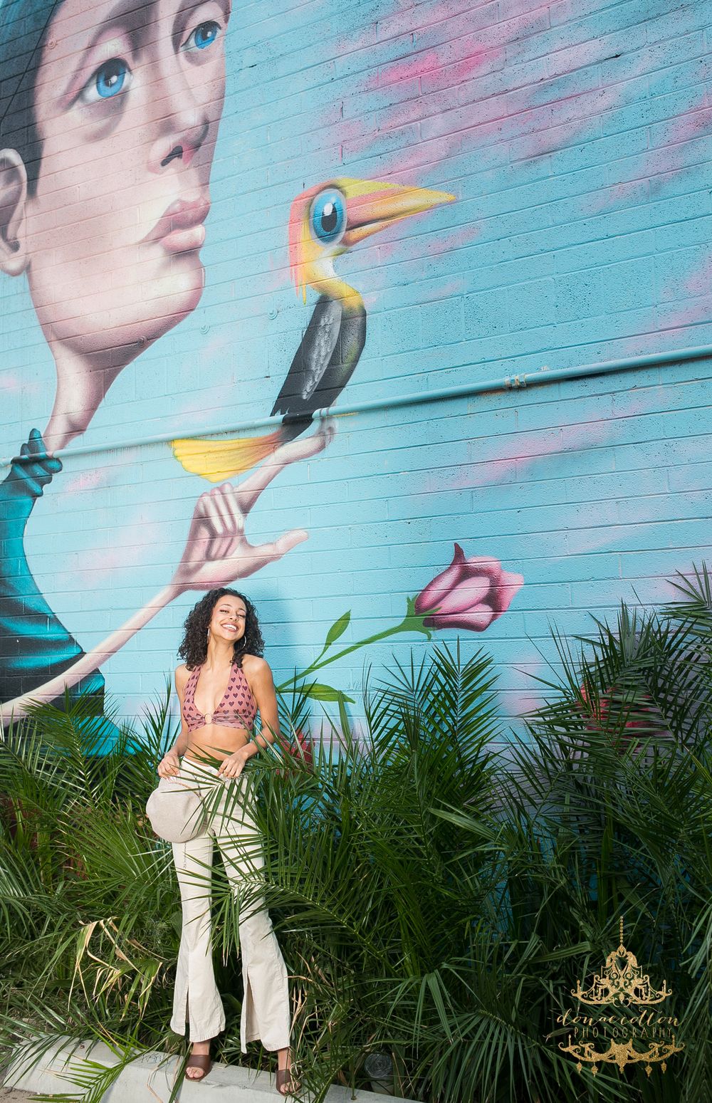 Woman in crop top and wide-leg pants poses before colorful tropical bird mural with palm plants.