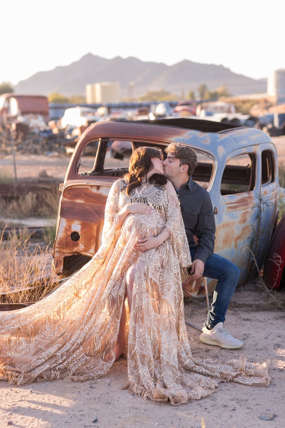 Couple kisses beside rustic vintage car at sunset in desert landscape, woman in flowing lace maternity gown.