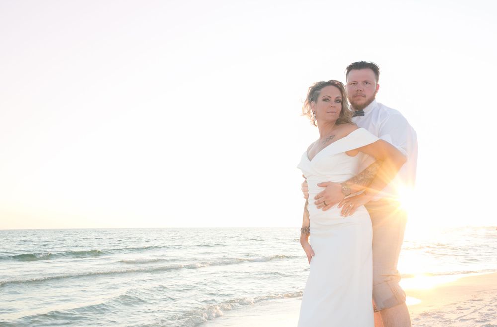 Couple embraces on beach at sunset, woman in white dress, golden backlight over ocean waves in Destin, FL