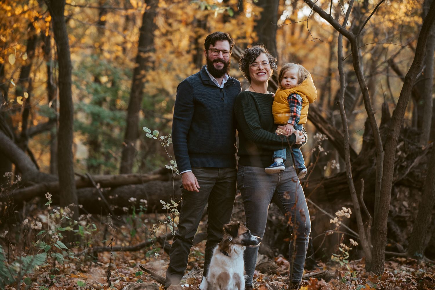 A young family with a toddler and dog poses in a colorful autumn forest setting.