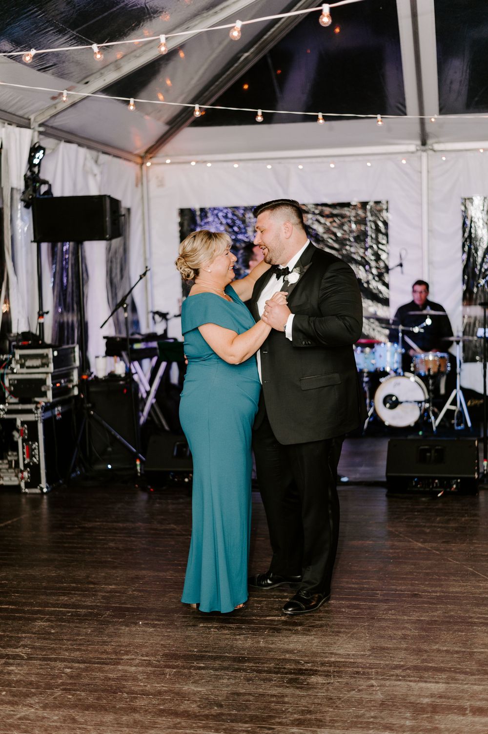A couple dances together on a wooden floor under string lights at a wedding reception.