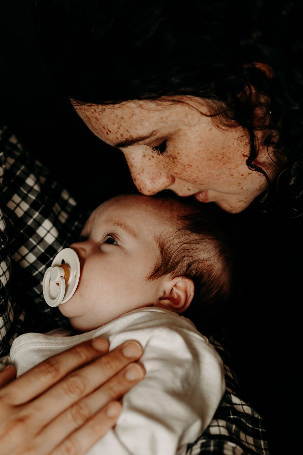 Intimate moment between caregiver and baby with pacifier in moody lighting.